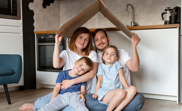 Family of four holding cardboard roof above their heads while sitting together on floor