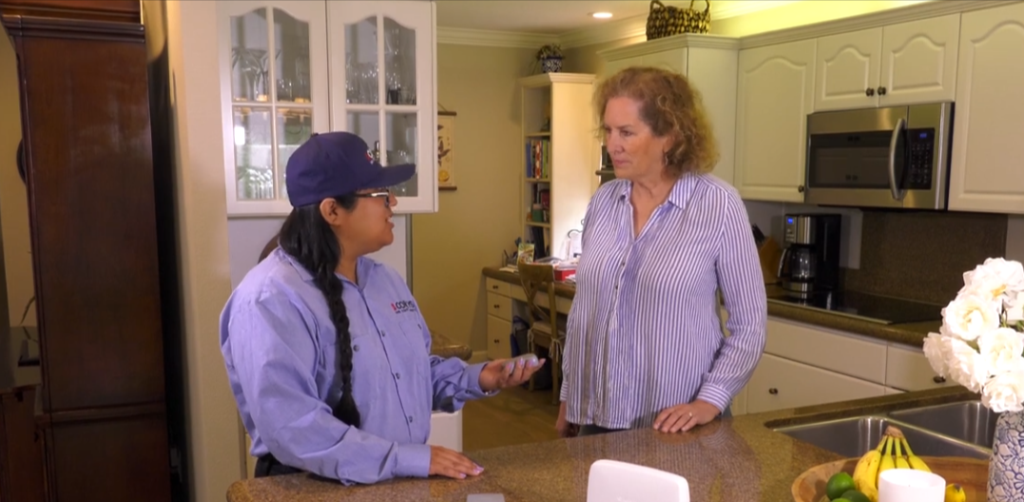 Two women conversing in a kitchen, one wearing a purple shirt and cap, the other in a striped button-up shirt