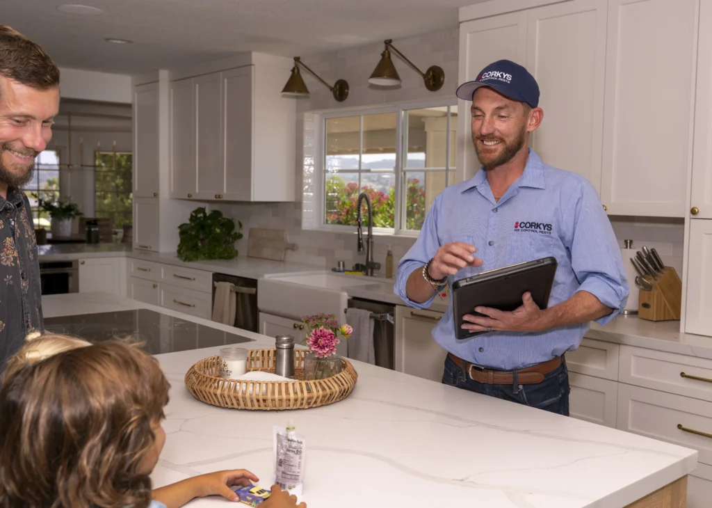 A pest control technician in a blue Corky's uniform holds a tablet while consulting with homeowners in a modern white kitchen