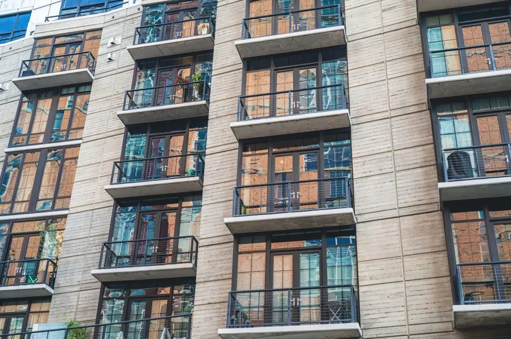 Modern residential building facade with brick and stone exterior, multiple balconies with metal railings, and large windows reflecting sunlight